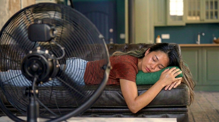 das Bild zeigt eine schwitzende Frau, die vor einem Ventilator auf einem Sofa liegt